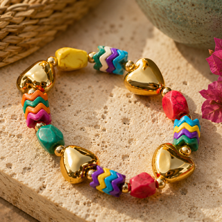A colorful beaded bracelet with shiny gold heart-shaped beads and alternating turquoise, pink, purple, and orange zigzag beads, displayed on a textured beige stone surface.