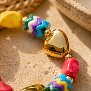 A close-up of a colorful beaded bracelet with a shiny gold heart charm, turquoise, purple, white, and yellow chevron beads, and pink stone beads, resting on a sandy surface.