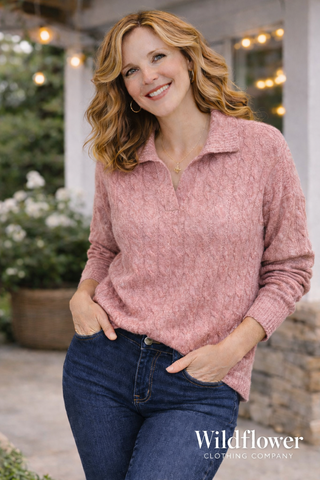 Woman wearing a pink sweater and blue jeans standing outdoors with 'Wildflower Clothing Company' branding.