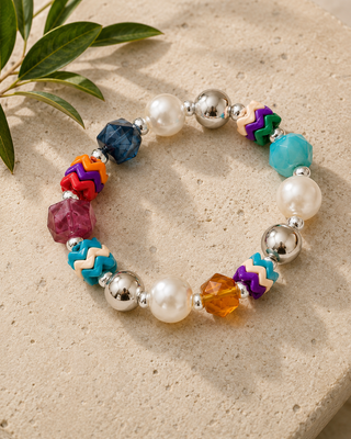 A colorful beaded bracelet with white pearl-like beads, silver-toned metal beads, and multicolor geometric and chevron beads, displayed on a textured beige stone surface with green leaves in the corner.