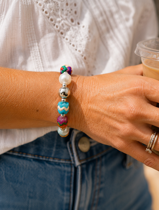 A close-up of a person's wrist wearing a colorful beaded bracelet with white pearl-like beads, silver-toned metal beads, and turquoise, pink, and purple accents. The person is wearing a white eyelet blouse and blue jeans, and is holding a clear-lidded iced coffee.