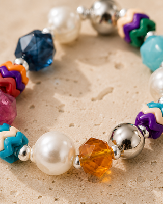 A close-up of a colorful beaded bracelet featuring white faux pearls, faceted crystal beads in blue, orange, pink, and amber, and chevron beads in purple, teal, orange, and cream, separated by shiny silver spacer beads, displayed on a textured beige stone surface.